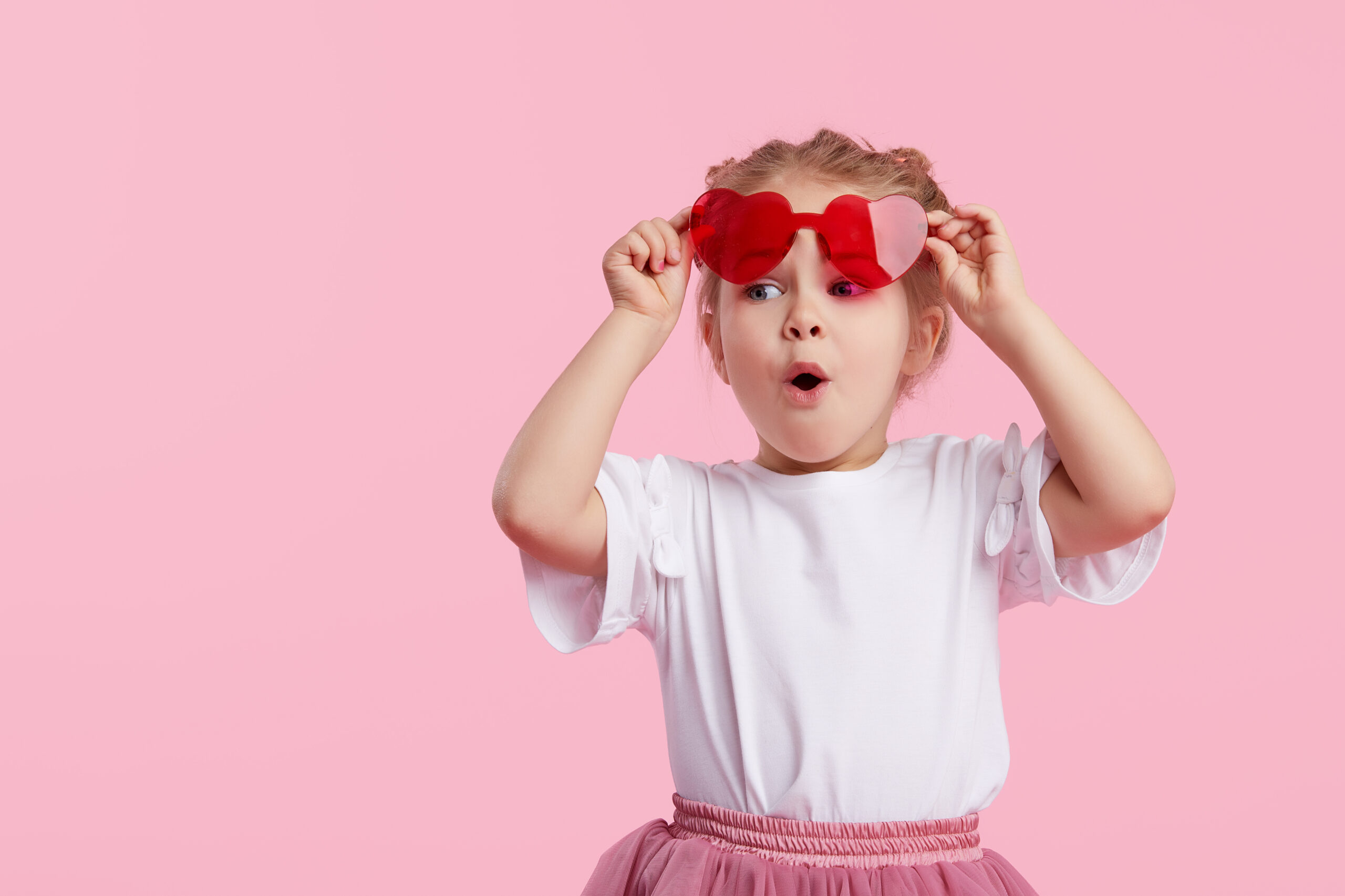 Portrait of surprised cute little toddler girl in the heart shape sunglasses. Child with open mouth having fun isolated over pink background. Looking at camera. Wow funny face