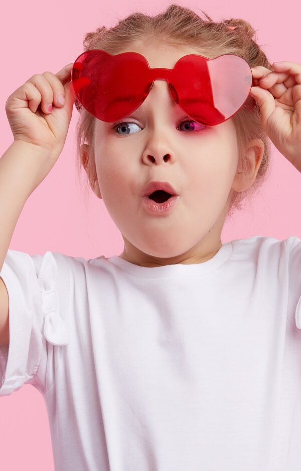 Portrait of surprised cute little toddler girl in the heart shape sunglasses. Child with open mouth having fun isolated over pink background. Looking at camera. Wow funny face